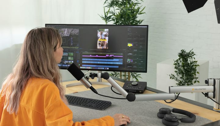 A woman sits at a desk with video editing software on the monitor and a boom arm with a mic.