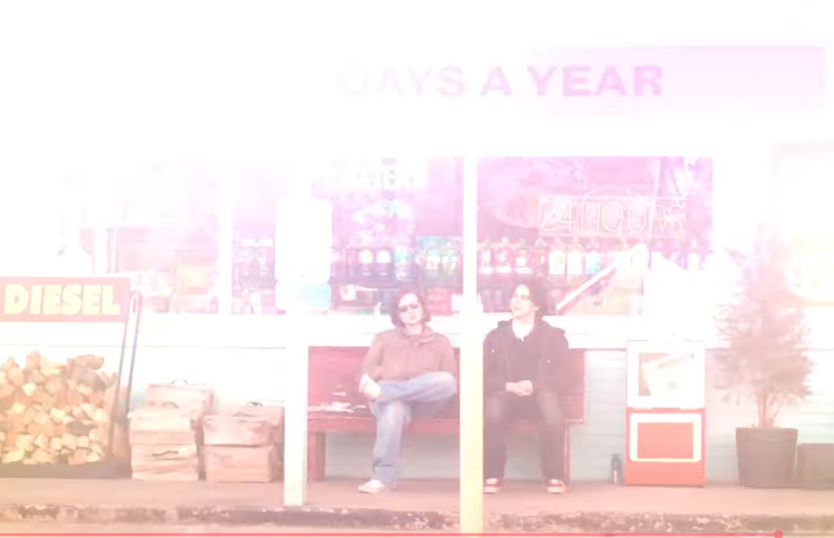 Screenshot showing two people sitting on a bench in front of a store from The Great Divide music video.