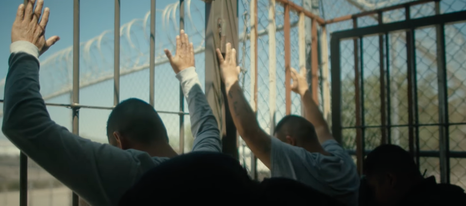 Screenshot from Narco Mennonites trailer showing two men up against bars with their hands raised being checked out by police